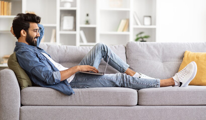 Young Smiling Indian Man Relaxing On Couch With Laptop