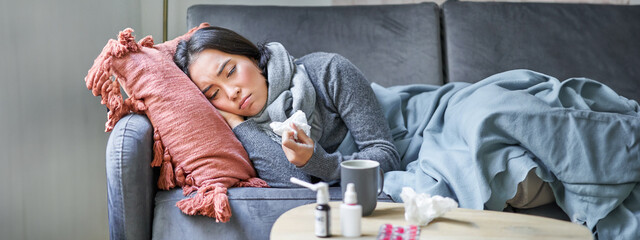 Sick sad korean woman lying on sofa, feeling unwell, catching cold, flu and temperature, looking upset, taking medication