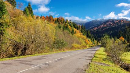 Fototapeta na wymiar straight road through mountainous countryside in autumn. trees on the hill in colorful foliage. grassy meadow on the roadside. warm sunny weather. old cracked asphalt