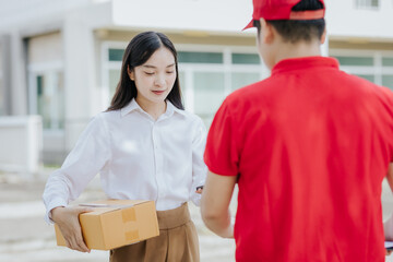 Young Asian woman receives a cardboard box from a smiling and happy delivery man at the front of...