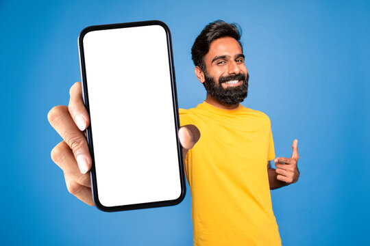 Online ad. Smiling middle aged indian man pointing at blank smartphone in his hand, blue background, mockup