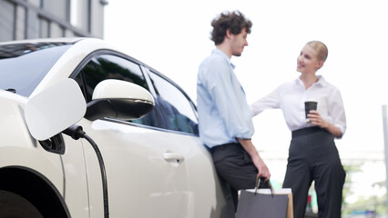 Progressive businessman and businesswoman leaning on electric car connected to charging station...