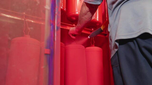 Worker spraying red paint on detail in painting chamber. Worker paints fire extinguishers with spray gun at factory. Close-up in 4K, UHD