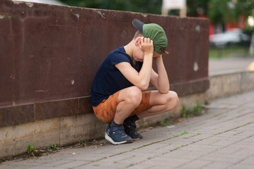 A teenage boy is squatting leaning against a concrete wall on the street. Upset and sad. Bullying....