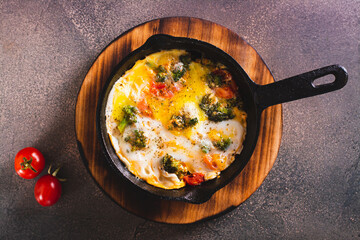 Close up of frying pan with scrambled eggs with broccoli and tomatoes on a wooden board top view