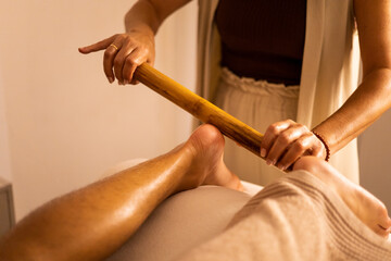 An unrecognizable beautician gives a man a wood therapy session on his feet in a beauty center. Use a wooden tool on the heel of the foot to massage. Biotherapy on the feet. Biotherapy concept - obrazy, fototapety, plakaty