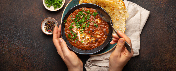 Female hands holding a bowl and eating traditional Indian Punjabi dish Dal makhani with lentils and beans served with naan flat bread, fresh cilantro on brown concrete rustic table top view. - obrazy, fototapety, plakaty