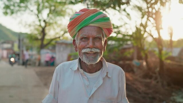 An Indian ethnic happy man or male is wearing a traditional turban or headwear facing or looking at the camera and smiling while standing outdoors in a village