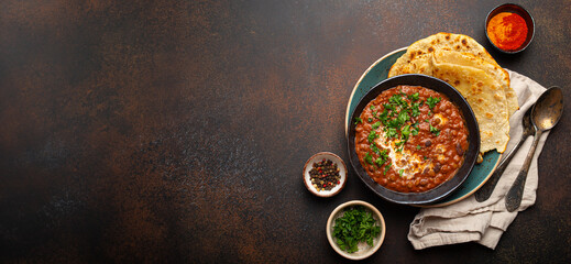 Traditional Indian Punjabi dish Dal makhani with lentils and beans in black bowl served with naan flat bread, fresh cilantro and two spoons on brown concrete rustic table top view. Space for text.