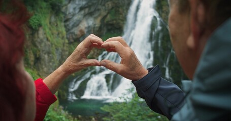 Older retired couple making heart with their hands while hiking to a waterfall in Austria. The...
