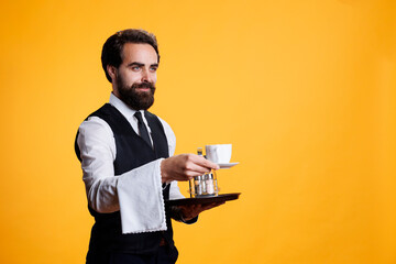Waiter giving cup of coffee to client on camera, pretending to serve people at fine dining restaurant. Skilled stylish man in formal attire having towel over arm and food platter.