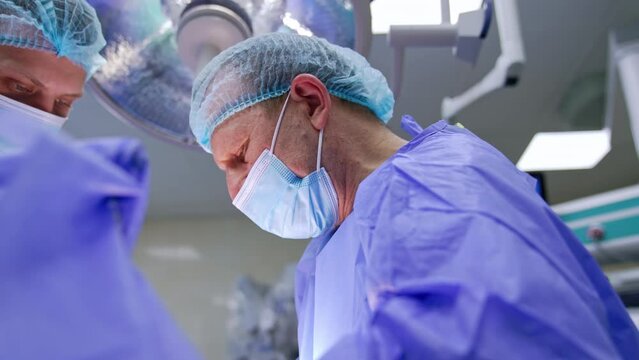 Old male experienced surgeon focused on operation. Younger doctor assisting the professional.