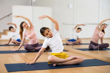 Fototapeten Positive sporty preteen girl doing stretching exercises while sitting in lotus position during training with family in fitness center © JackF