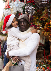 Joyful man holding small daughter, looking around at Christmas fair..