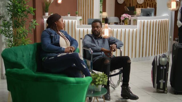 Wheelchair user hotel client in lobby, african american man suffering from chronic disability and going on holiday. Young couple at reception travelling despite reduced mobility causes.