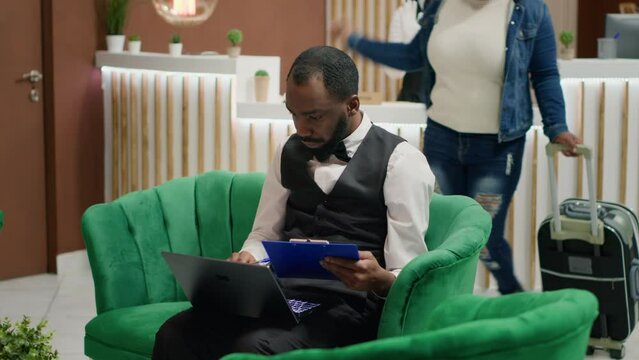 Tourism industry employee. Bellboy entering data document forms on papers in lounge area, hotel concierge making entry in daily luggage movement register, using clipboard notes.