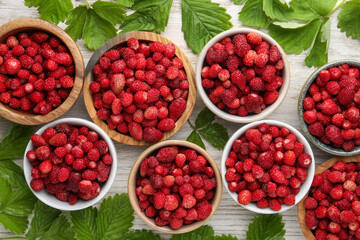 Fresh wild strawberries in bowls and leaves on white wooden table, flat lay