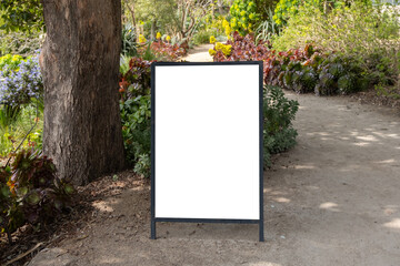 Blank white mockup background template of a standing board placed outdoor on a gravel footpath surrounded by beautiful garden plants. Empty information penal for wedding or events in countryside.