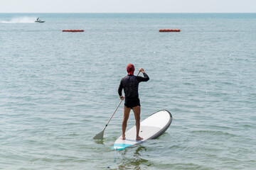 A man stands on a SUP board that floats on a calm sea and rows with an oar. Quiet sunny weather at a tropical resort.