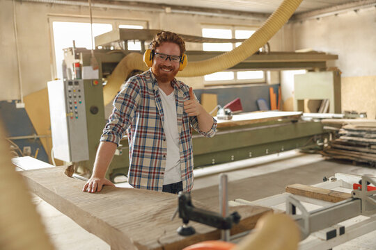 Smiling carpenter cutting wooden board at sawmill carpentry manufacturing and showing thumb up