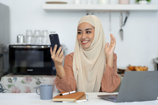 Beautiful Asian Muslim businesswoman or entrepreneur wearing hijab working on holiday at home using her phone for communication. Video calls via social media apps happily. lifestyle concept