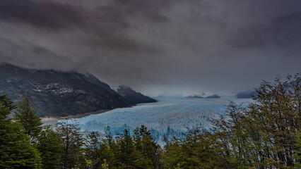 The impressive Perito Moreno Glacier stretches to the horizon between the mountains. A mass of blue ice with sharp peaks, cracks, crevices. Cloudy. Green vegetation in the foreground. El Calafate. 