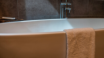 Details of the bathroom interior. A terry towel is draped over the edge of a large white acrylic bathtub. Close-up. Metal taps on the beige tiled wall. - obrazy, fototapety, plakaty