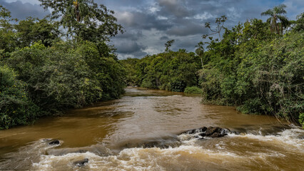 The Iguazu River flows in the jungle between banks overgrown with lush tropical vegetation. The water boils and foams on the rocks. Clouds in the sky. Argentina. Iguazu National Park.