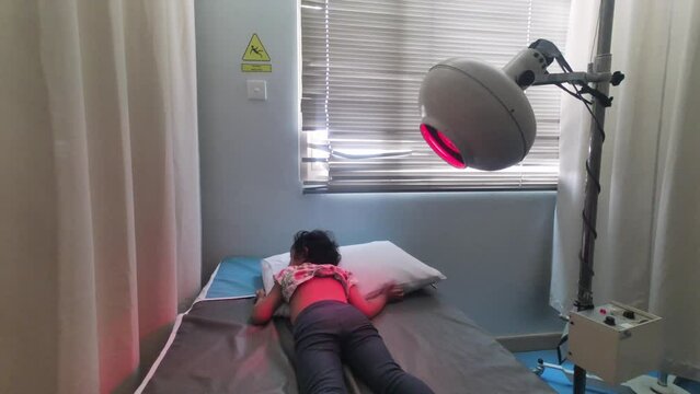 An Asian little girl lying in the bed with a heat lamp on her back in the hospital. Infrared heating therapy.