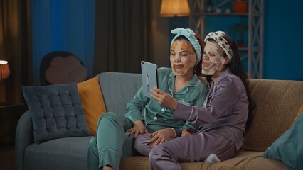 Medium-full photo capturing two young women in pajamas sitting on the couch and doing their evening skincare routine, wearing face masks, having a video call on a tablet.