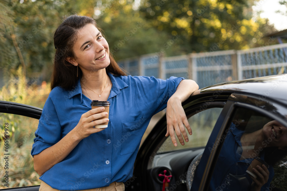 Wall mural Business lady standing with a glass of takeaway coffee near her car with the door open outdoors