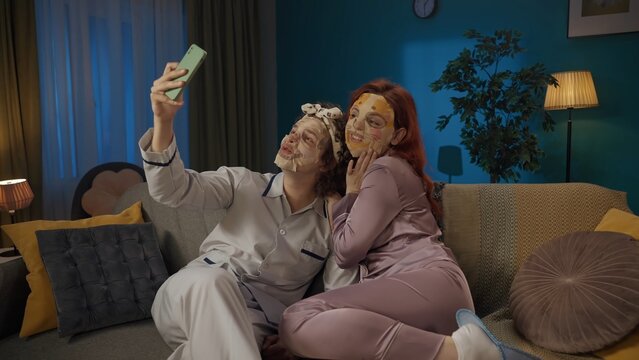 Portrait of young couple spending time together. Man and woman sitting on the sofa, wearing face masks, taking selfie on smartphone.