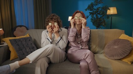 Portrait of young couple. Man and woman sitting on the sofa holding round slices of cucumber over eyes, smiling at the camera.