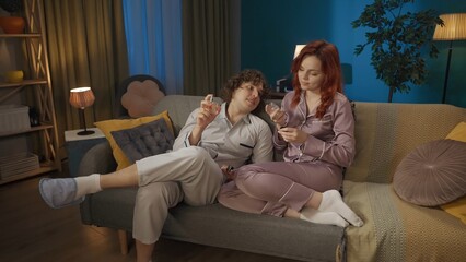 Obraz premium Portrait of young couple spending time together. Man and woman in pajamas sitting on the sofa, holding perfume bottles.