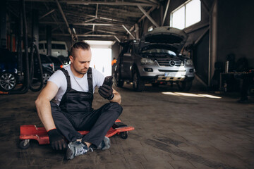 Young professional technician car mechanic man 20s in blue overalls white t-shirt use hold mobile cell phone browsing fix problem with raised hood bonnet work in vehicle repair shop workshop indoor.