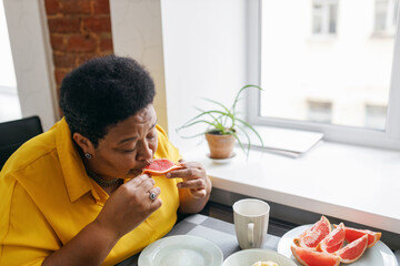 Upper side view image of plus size african american senior retired woman eating juicy fresh organic grapefruit sitting at table near window, being on diet, loosing weight to be healthy