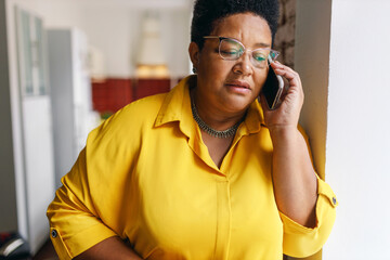 Serious african american senior woman in glasses and yellow shirt talking on phone with serious worried facial expression, receiving bad news from her doctor or relatives, leaning on wall - 655584998