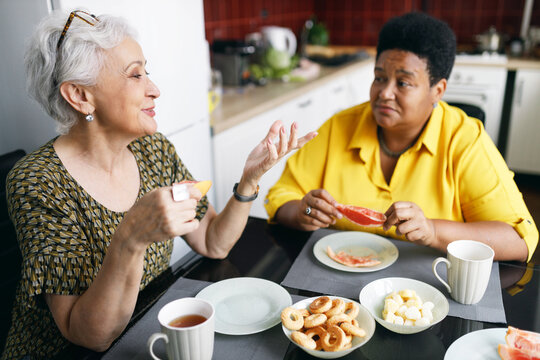Side view image with selective focus on elderly charming caucasian female gesticulating and smiling while talking to her african american female friend while drinking tea together at kitchen table