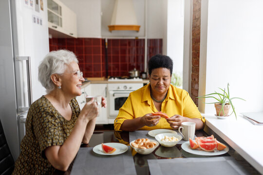 Side view indoor image of cute lovely retired female friends of diverse ethnicity spending time together sitting at kitchen having nice conversation while drinking tea with fruits and sweets