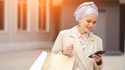 Muslim businesswoman types messages to friend about shopping day. Woman with excited expression shares emotions returning from shopping mall, sunlight