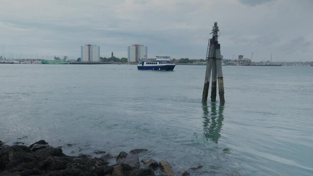 Wide shot of seawater hitting rocks and post on a calm day in the harbour, a boat is passing by with council estates in the background. Handheld shot. 1080p.