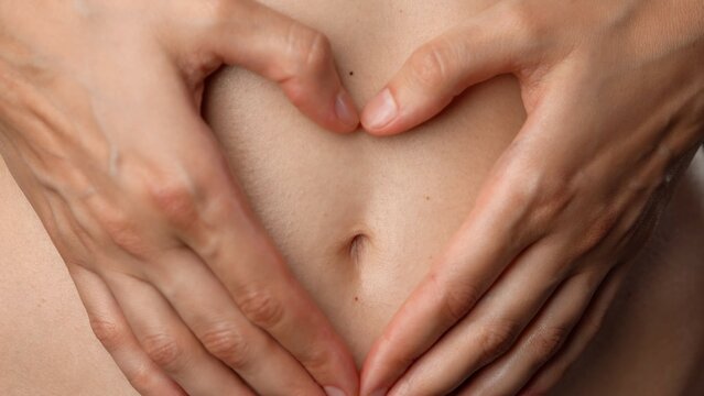Closeup shot of attractive caucasian woman body part, touching tummy area with hands in heart shape, smooth natural skin.