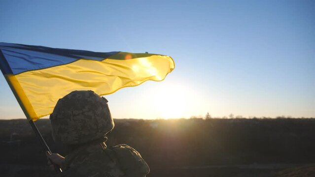 Close up of ukrainian army man stands at hill waving blue-yellow banner against sunset. Male soldier in military uniform lifting flag of Ukraine as a symbol of victory against russian aggression