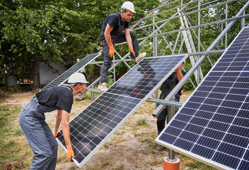 Three men solar installers wearing safety helmets and work overalls while mounting photovoltaic solar panel system. Male workers installing solar module on metal construction.