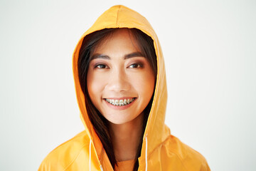 Close up portrait of joyful asian girl with braces dressed in bright yellow raincoat posing with hood on her head