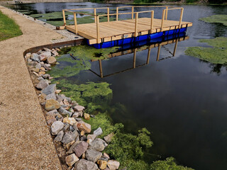 Pier for park visitors. two steps lead to it. planks terrace over water. Pond with a dock for boats. floor plan of a rectangle. the terrace has no railings., stone, green, algae, boat, duckweed, lawn