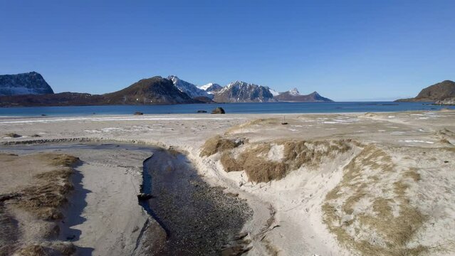 Aerial dolly forward shot skimming over the sand dunes at famous Haukland beach in Lofoten Norway in late winter with small patches of snow and stunning mountain views over the water