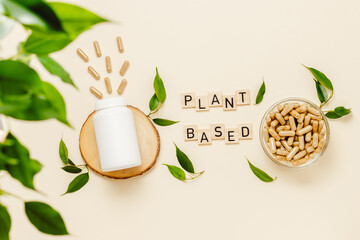Medical white bottle mockup on wooden podium with pills and wooden letters, plant based text, green leaves. Organic medication, natural herbal supplement, bio vitamins, top view