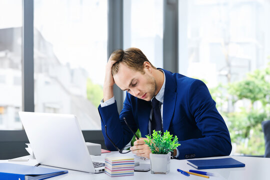 Stressed, tired businessman or heaving headache, hold his head, in blue suit, work with laptop at office. Business fall, job or education concept. Executive employee sitting near window on background.