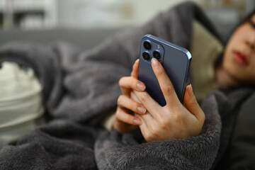 Upset ill woman young woman in blanket lying on couch and using smartphone for searching medical information on internet